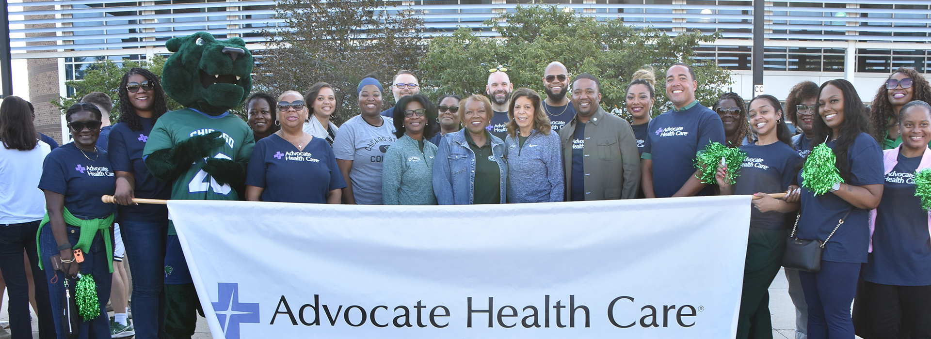 Leadership of Chicago State University and Advocate Health Care smile in front of the Jones Convocation Center, holding a wide white banner that reads "Advocate Health Care." A green cougar mascot in a sports jersey stands on the left side of the group, and several attendees hold green pompoms.