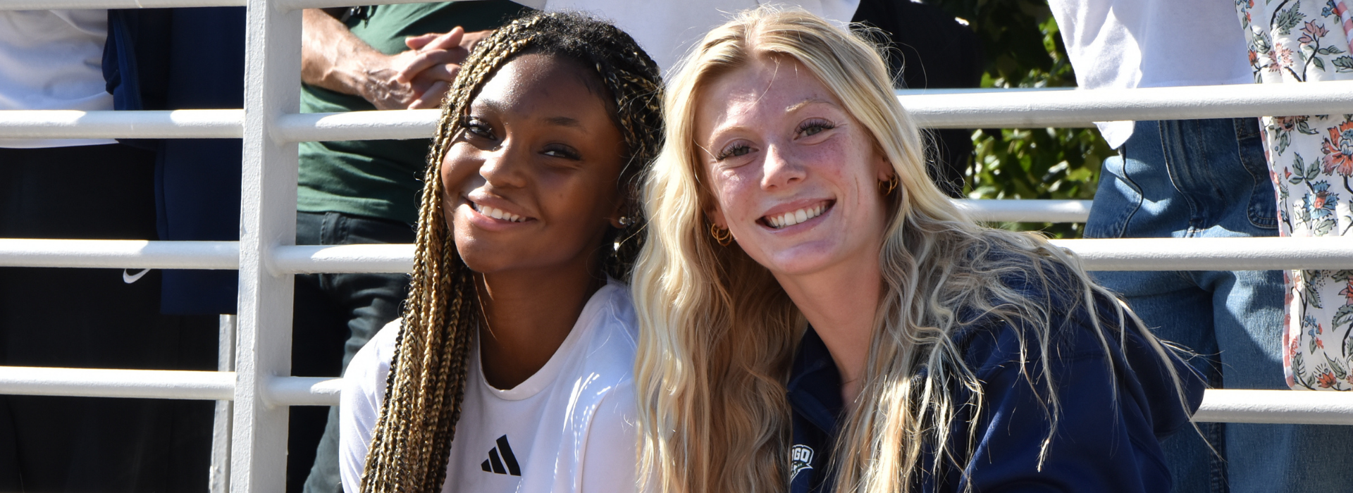 Two young women smile while sitting in bleachers at an outdoor campus event. The student on the left has long braided hair and wears a white Adidas shirt, and the student on the right has long blonde hair and wears a navy blue Chicago State athletics jacket.