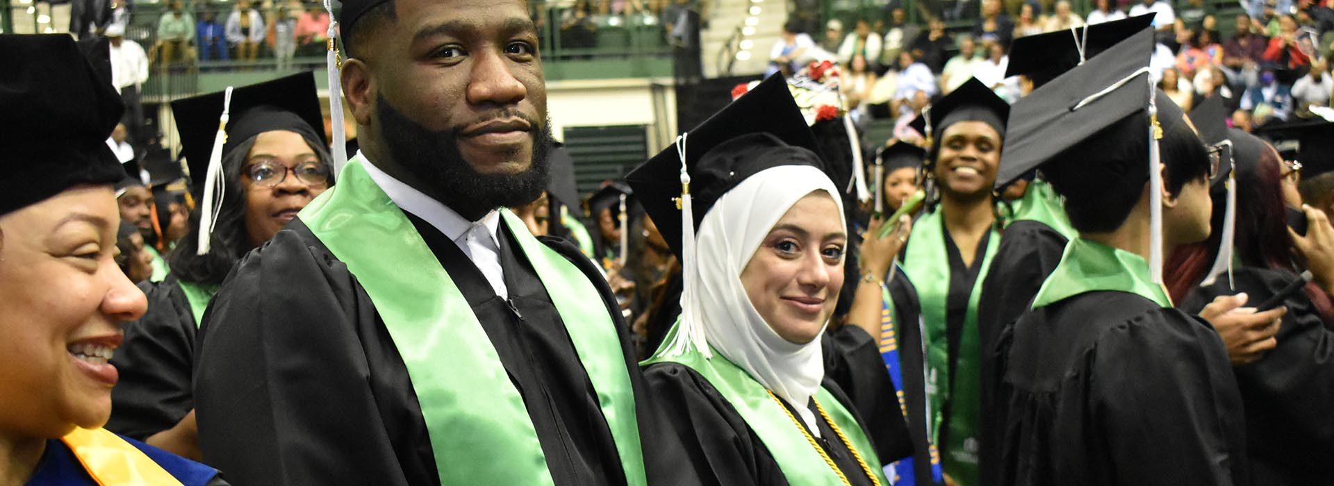 A diverse group of Chicago State University graduates in black caps and gowns with green and yellow stoles smile during commencement ceremony, with a packed arena visible in the background.