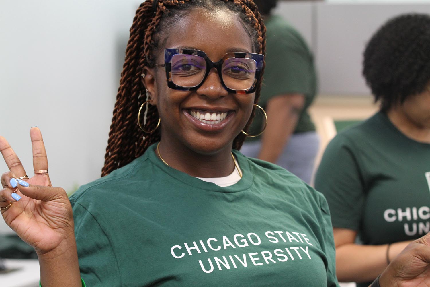 Close-up photo of a student smiling and making a peace sign with her hand inside a classroom. She's wearing a green Chicago State University t-shirt, and dark-brown rimmed glasses.