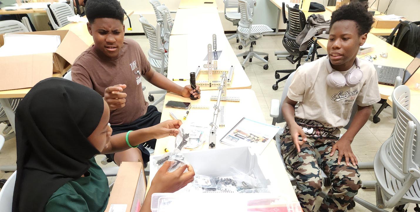 Three students collaborate around a table in a classroom while assembling parts from a robotics kit.