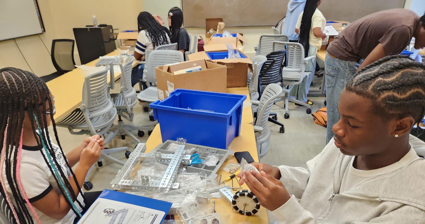 Two students sit across a lab table, unbagging small hardware components to assemble a robotics frame.
