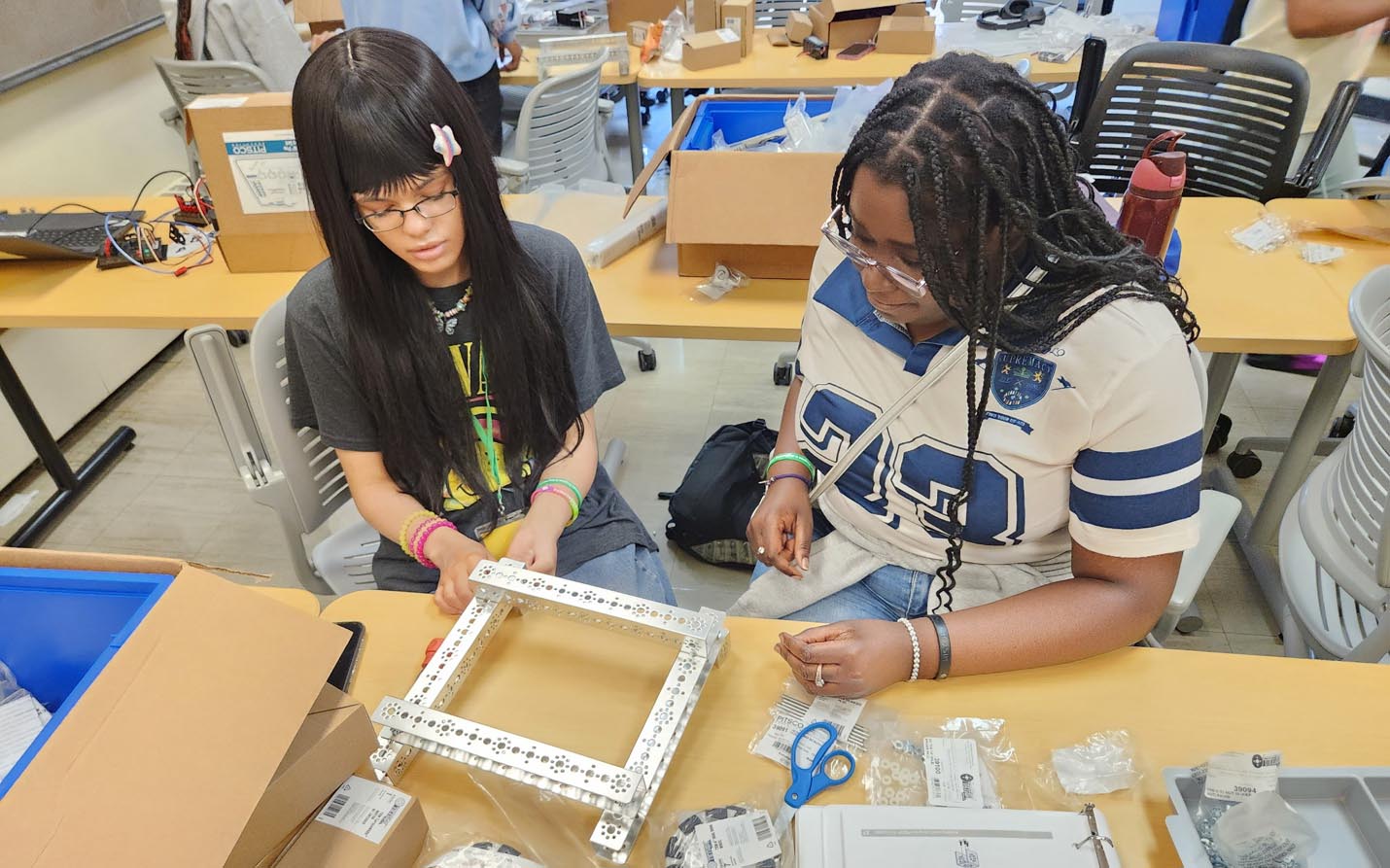 Two students collaborate at a desk to construct a square metal robot frame using parts and scissors from an engineering kit.