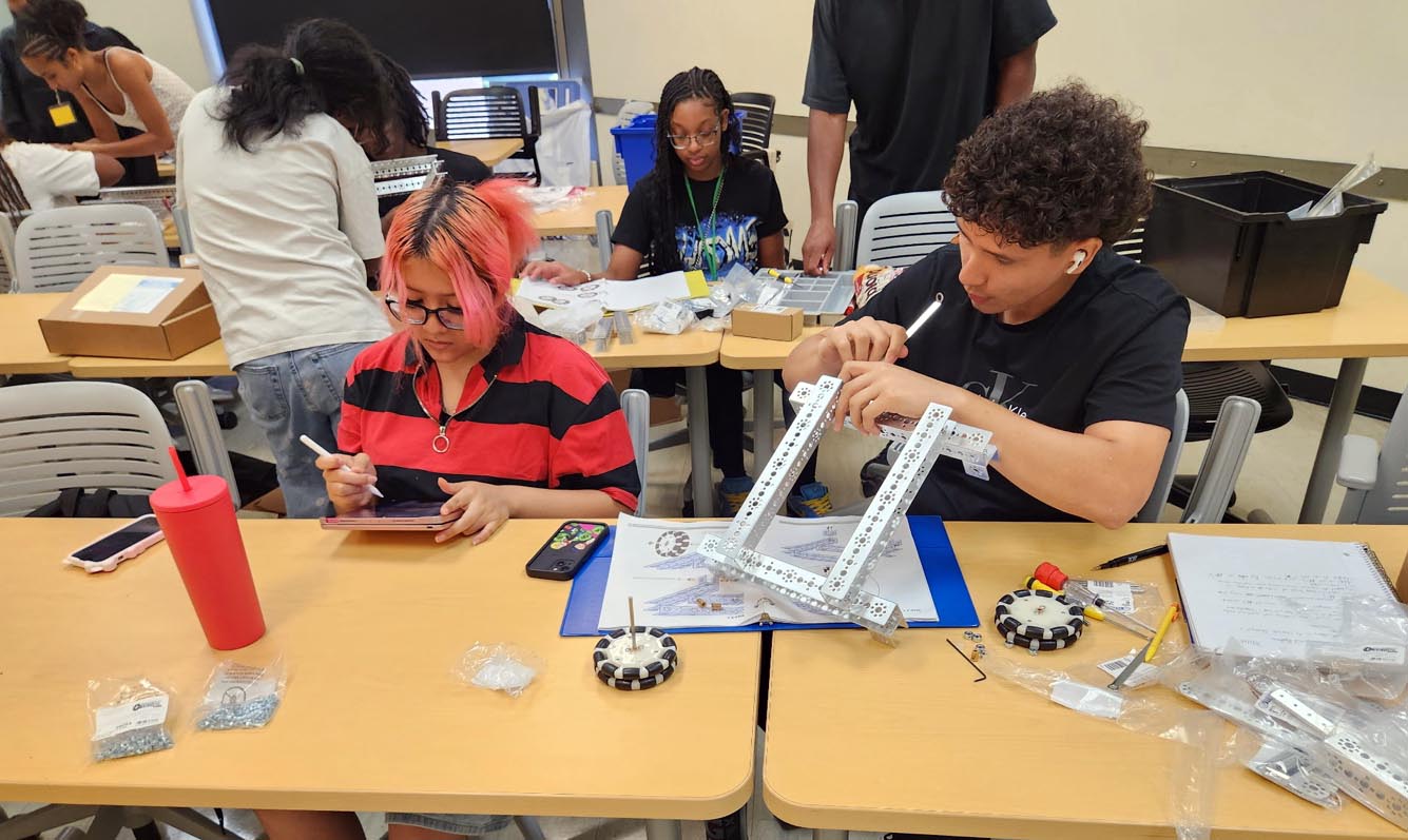 Two students sit at a classroom desk—one reviewing notes on a tablet with a stylus while the other uses a hand tool to assemble a metal robotics frame.