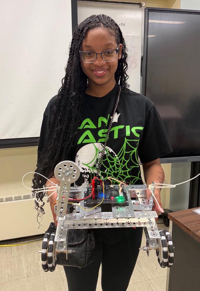 A young woman wearing glasses smiles while holding up a finished four-wheeled metal robot prototype inside a classroom.