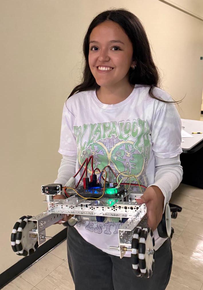 Another young woman smiles while holding up her finished four-wheeled metal robot prototype.