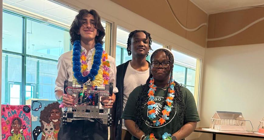 Three students posing together with floral leis, with one student holding up a completed custom robot prototype.