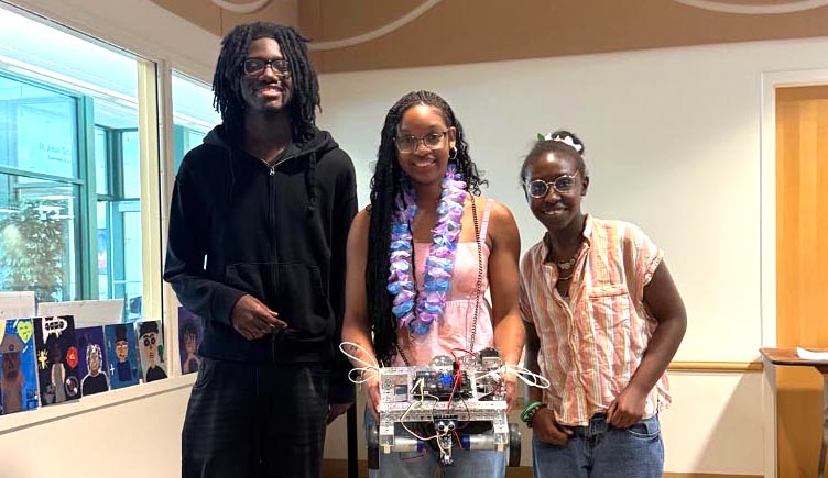 Three smiling students pose together, with the middle student wearing a purple floral lei and holding a finished custom robot build.