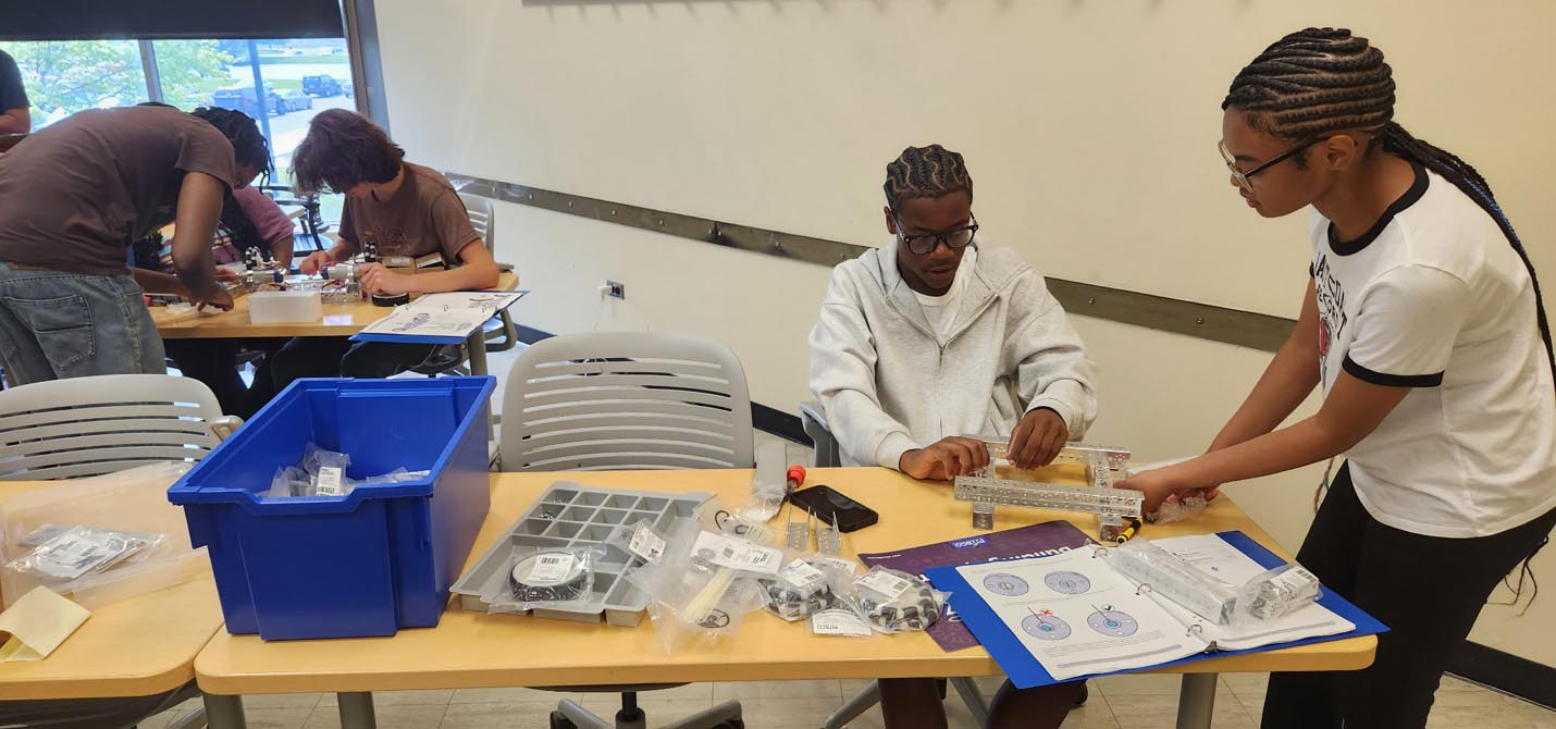 Two students assemble a metal robotics chassis at a classroom work table filled with parts, tools, and instruction guides.