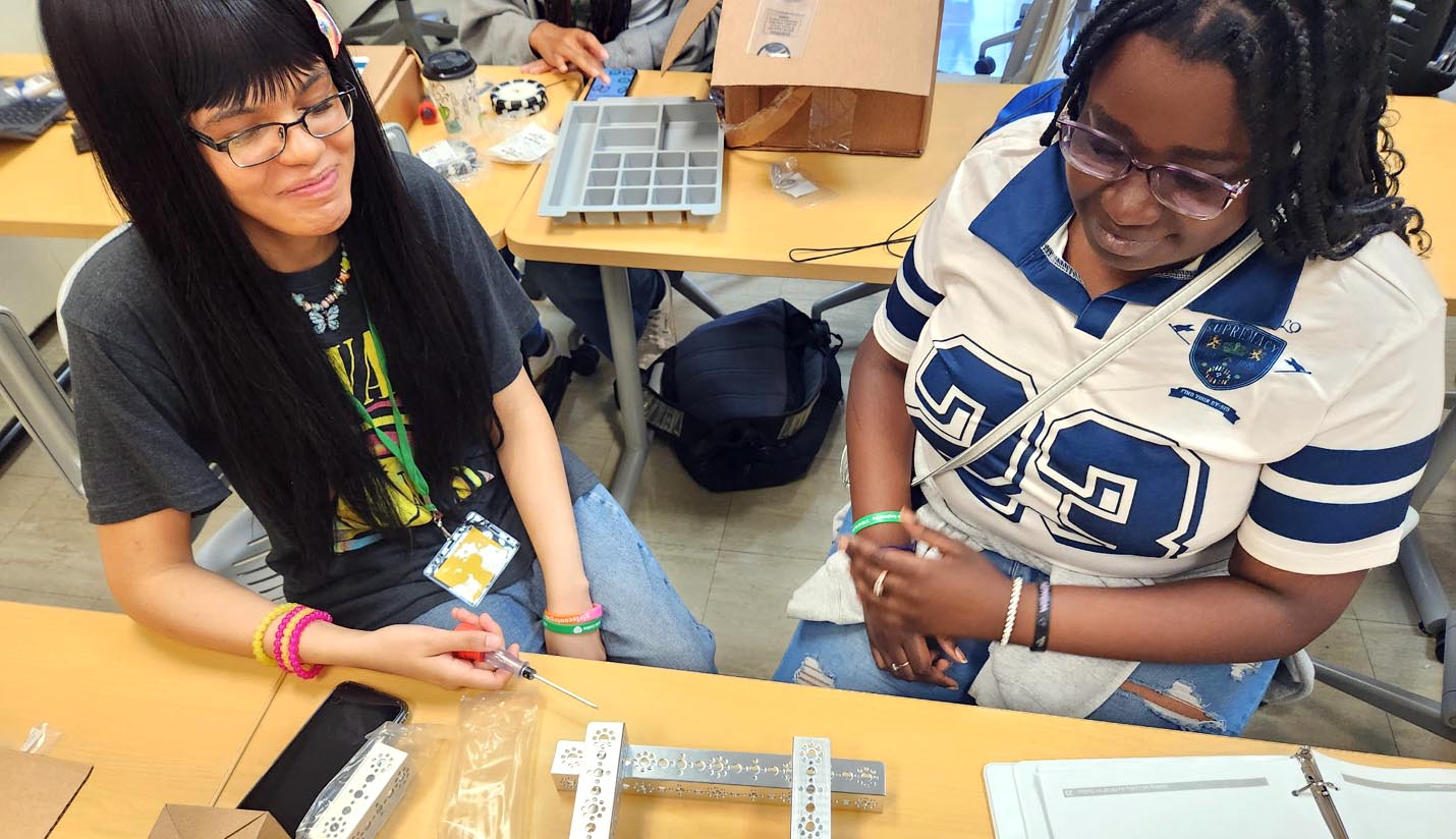 Two students work together at a desk, using a screwdriver to assemble a metal robotics component during a hands-on lab activity.