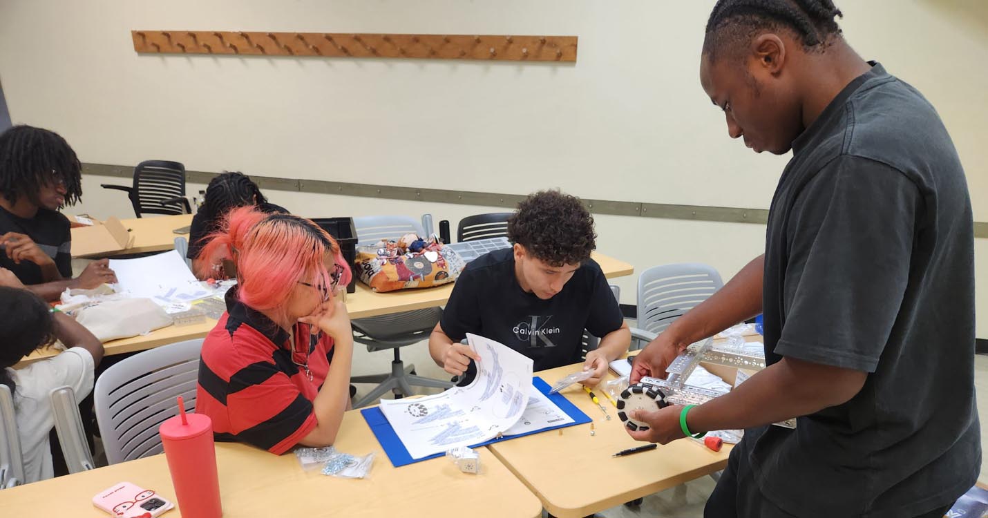 A student standing at a table demonstrates a robotic wheel component to seated classmates reviewing assembly instructions.