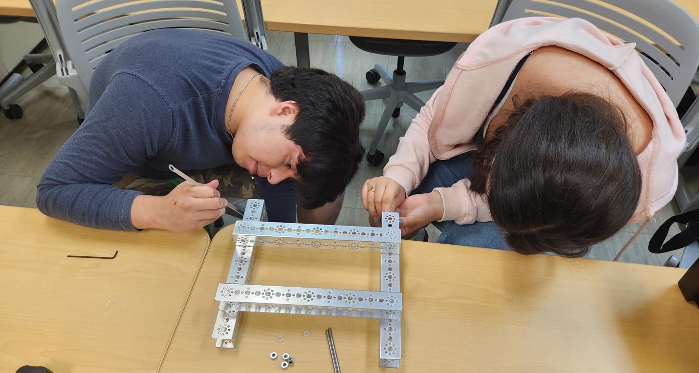 Two students lean over a table to carefully assemble a rectangular metal robotics frame using small hand tools.