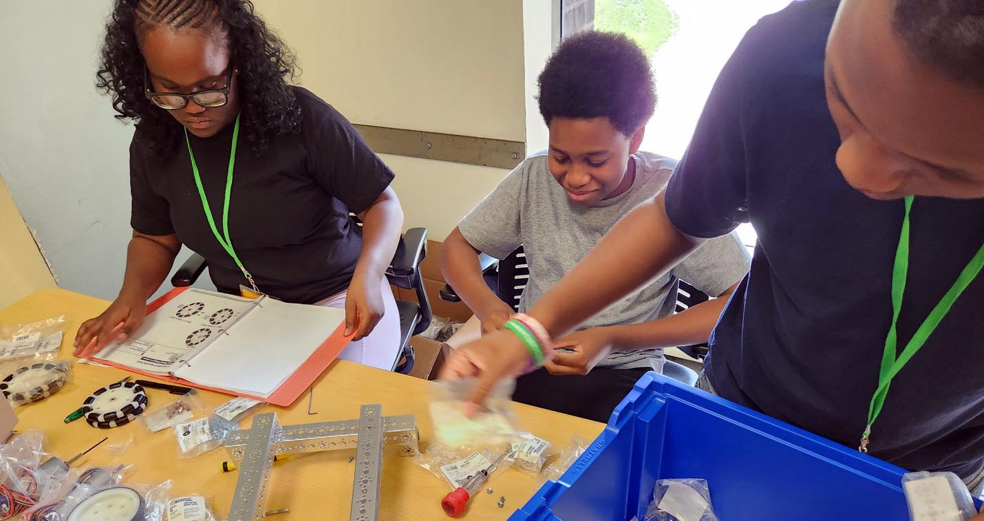 Three students wearing green lanyards work around a table, reviewing an instruction binder and selecting robotics parts from a blue storage bin.