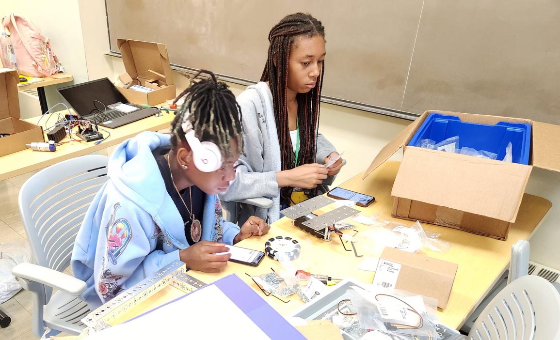Two young women sit at a lab table filled with robotics parts, tools, and cardboard boxes. One student wearing headphones checks instructions on a smartphone while her classmate holds small component packaging.