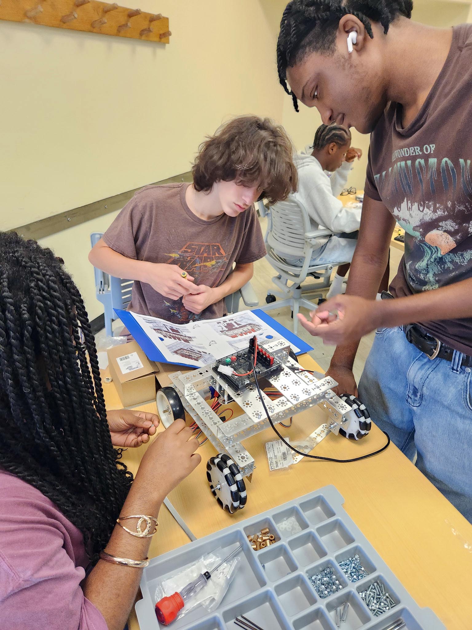 Three students collaborate around a work table to assemble a four-wheeled metal robot using printed diagrams and small tools.