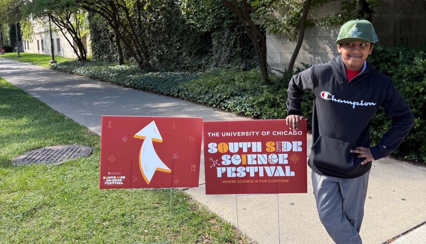 A young boy wearing a green Chicago State hat and a black hoodie leans against a University of Chicago yard sign for the South Side Science Festival.