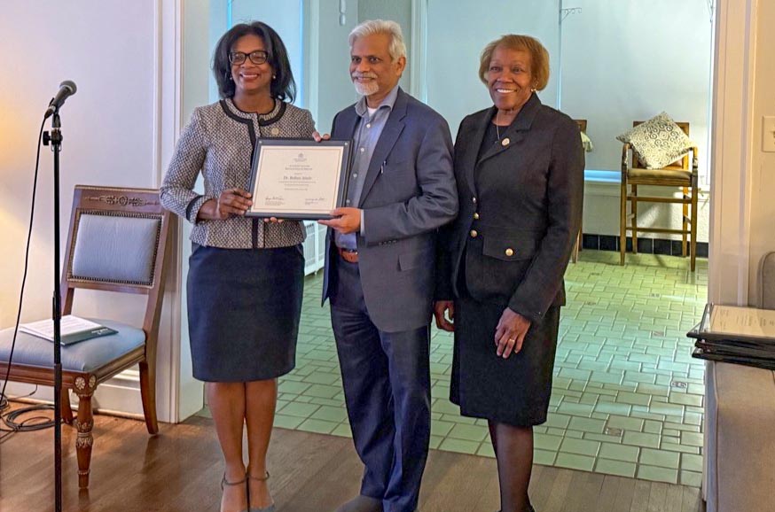 Dr. Attele accepts a certificate of appreciation from Provost Sonja Feist-Price and President Scott during a retirement celebration. On the left is a microphone and an ornate antique chair.