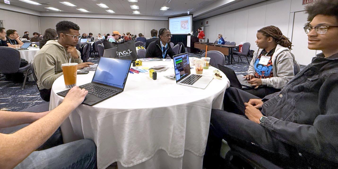 Five students seated at a round table in a large conference room with their laptops open.