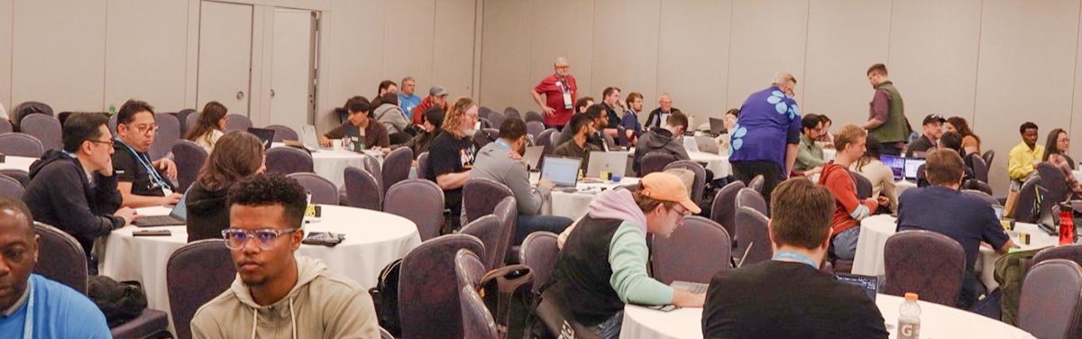 A large group of people sitting at round tables in a conference room, many working on laptops and engaging in discussions.