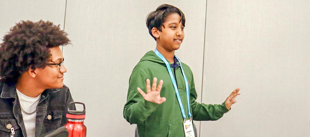 A young person in a green hoodie stands and gestures while speaking during a presentation or workshop, while another person in a denim jacket listens nearby.