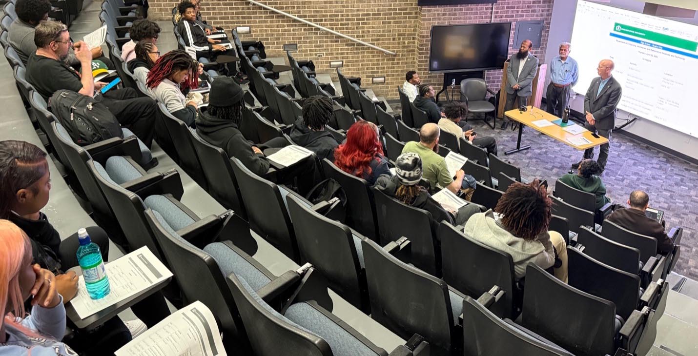 A group of high school students sitting in a theatre-style classroom listen intently as faculty members from the department of Computing, Information and Mathematical Sciences, and Technology (CIMST) speak. 