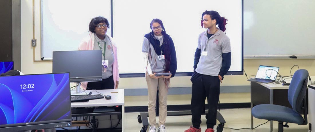 Three high school students wearing gray polo shirts stand in front of a computer lab.