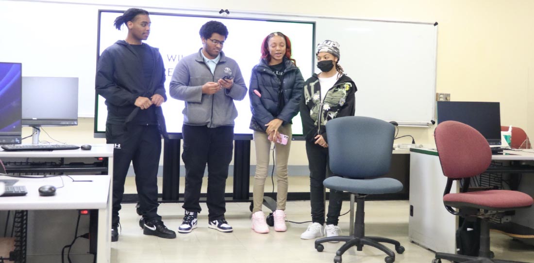 Four high school students standing at the front of the computer lab in front of a brightly lit smart board.