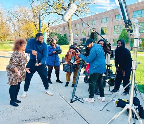 A CSU student film crew and cast film an outdoor scene on a campus sidewalk using a professional camera, boom microphone, and lighting gear.