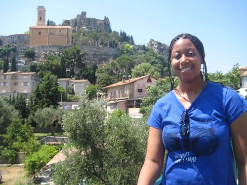 A woman smiling outdoors on a bright day with a hilltop village and historic church rising in the background.