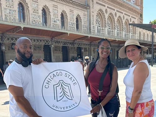 Two students stand alongside their advisor in a Spanish corridor holding a Chicago State University flag.