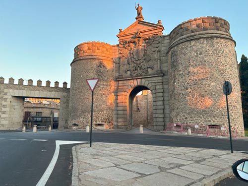 A large historic stone gate structure flanked by two rounded towers under warm sunlight, with an arched entryway leading into a city street.