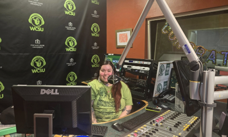 A woman sitting in the WCSU radio station control room smiling from behind a desktop computer monitor and mixing console.