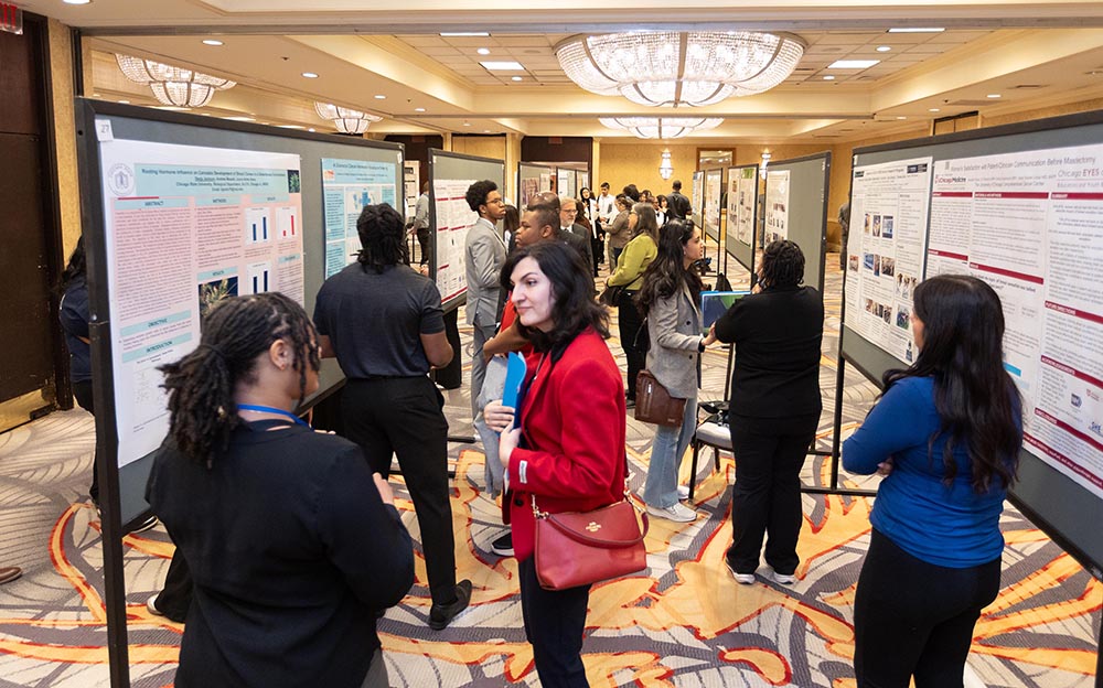 Many people are gathered in a conference hall, looking at large research posters displayed on tall easels throughout the room. The room is well-lit by chandeliers, and attendees are interacting and discussing the content of the posters.