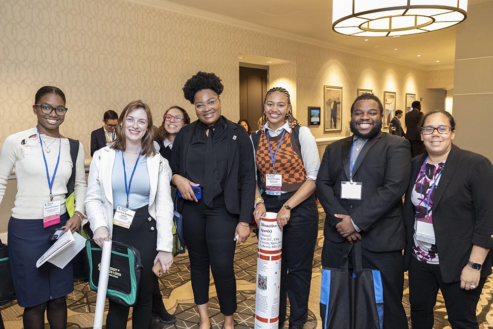 Six professionally dressed student researchers stand next to each other and pose for a photo. They’re in a warmly lit conference hall with a chandelier above them. In the background are more conference attendees and a display of four art canvases hanging on a hallway wall.