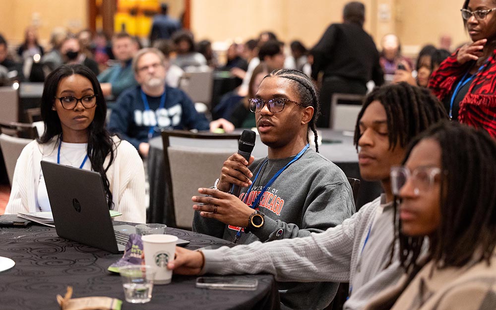 Four students sit at a round table covered in a black tablecloth. One of the students is speaking into a microphone