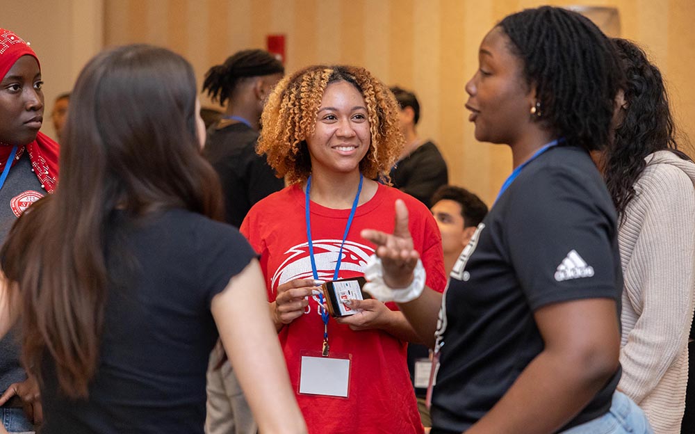 A close-up photo of three women standing in a circle talking while others converse in the background.