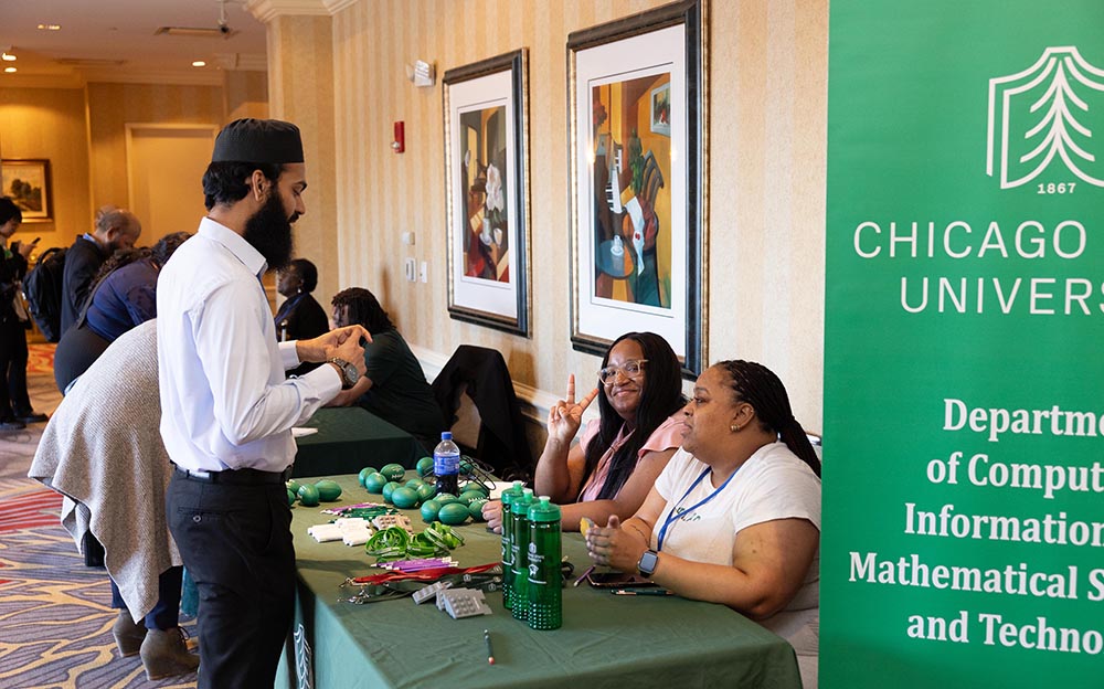 A man stands in front of a table chaired by two people. The table is covered with a green tablecloth and has swag items including water bottles, lanyards and small, soft footballs.