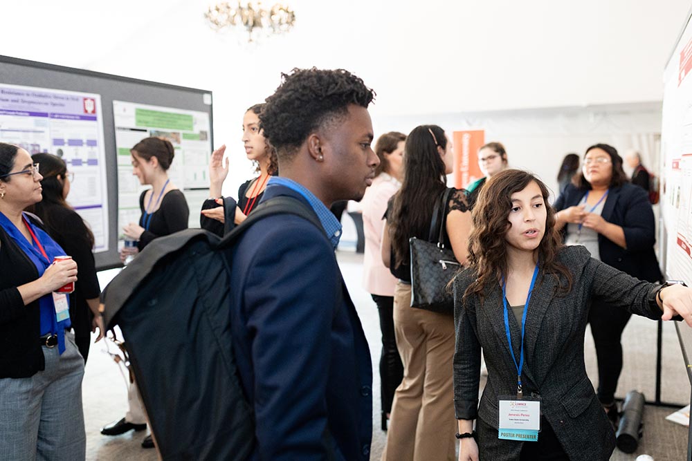 A vibrant conference scene showing a poster presentation session. A diverse group of attendees is views and discusses research posters. In the foreground, a woman with long, curly hair is presenting her poster to a man in a dark suit and backpack.