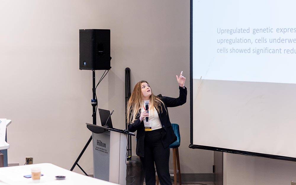 A man in a yellow hoodie and a navy-blue blazer talks into a microphone while sitting at a table during a panel discussion with three other people.