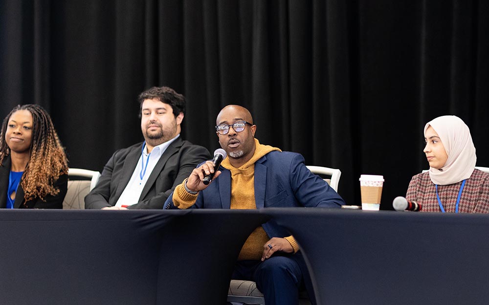 A man in a yellow hoodie and a navy-blue blazer talks into a microphone while sitting at a table during a panel discussion with three other people.