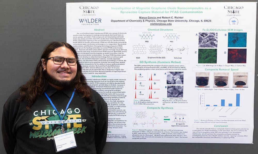 A student researcher with long, dark, wavy hair smiles and stands in front of his research poster. He’s wearing glasses, a black hoodie that says Chicago State Welcome Week and a blue conference lanyard.