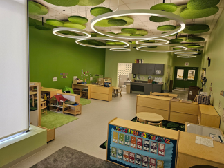 A pre-k classroom with bright green walls and modern, ring-shaped lighting suspended from the ceiling. Low, wooden shelves include toys and books, and there are activity areas for creative play. A handwashing station can be seen in the background