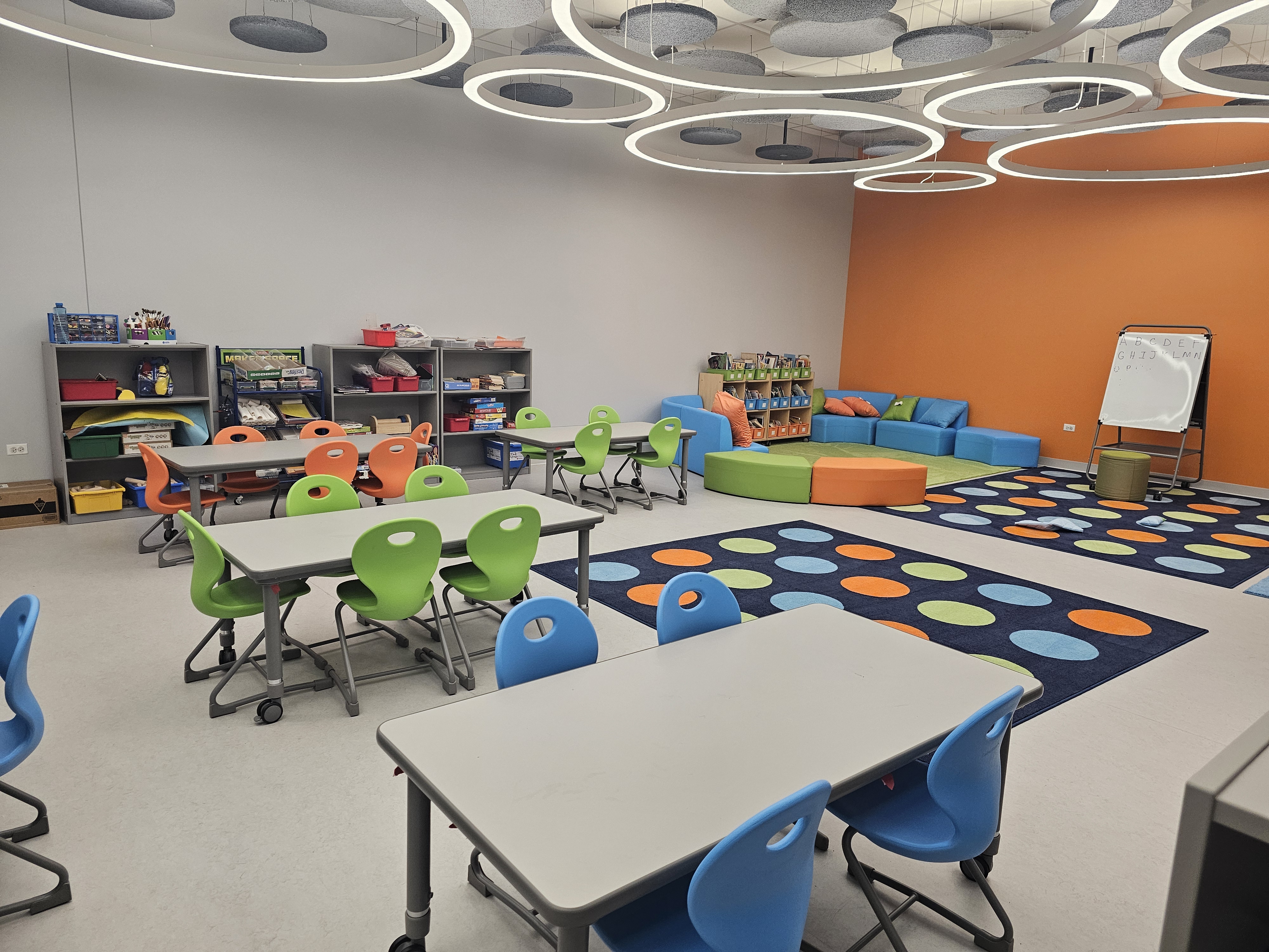 A bright and modern school-age classroom. The room is decorated with a variety of colorful furniture, including shelves filled with books, small tables with green chairs and blue sofas. Several rugs with large, multi-colored polka dots are on the floor. The ceiling features a unique lighting design with circular, suspended fixtures.