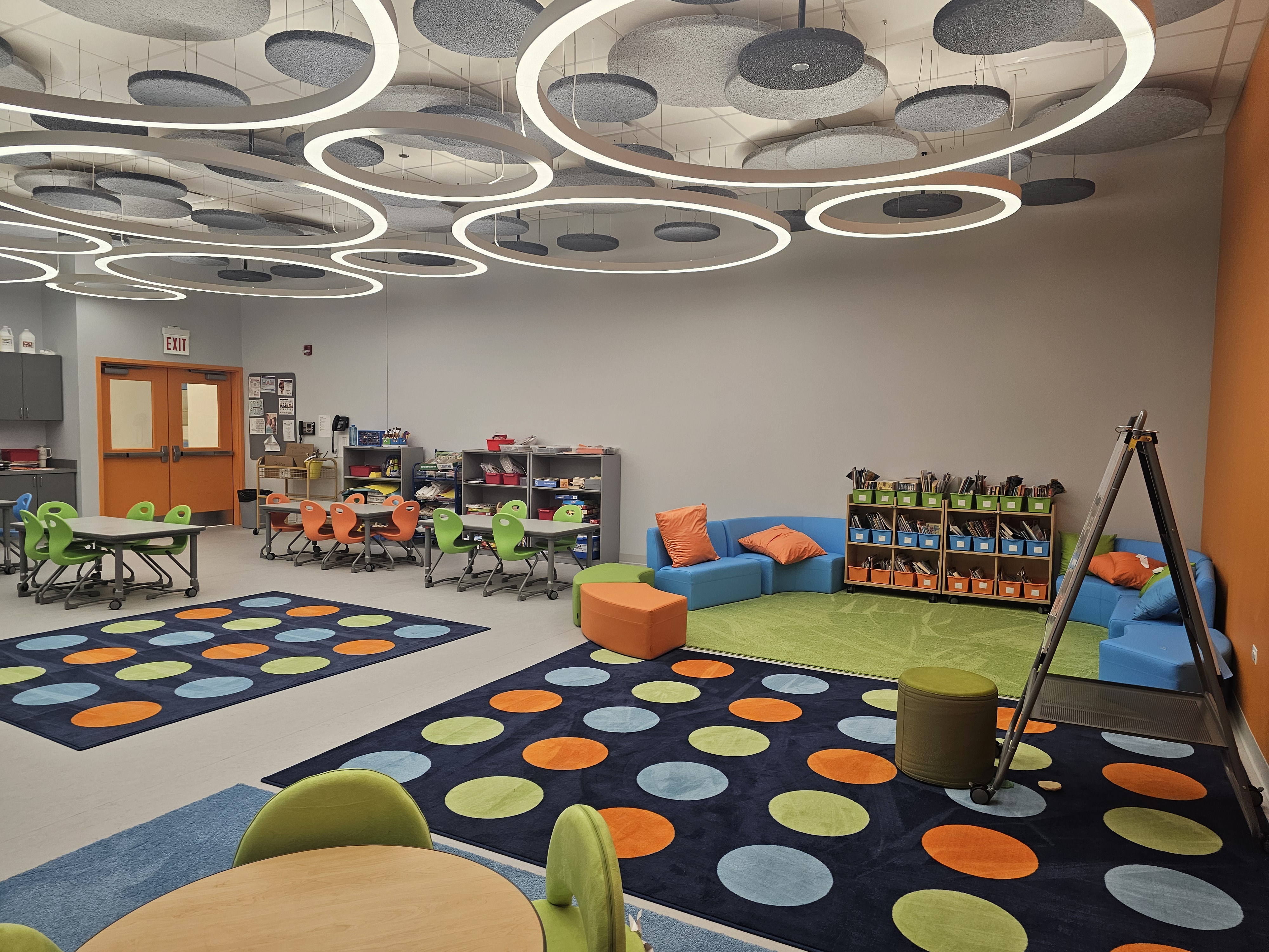 A spacious and colorful school-age classroom. The room is furnished with several small tables and chairs in the colors green, orange, and blue. A cozy corner with blue sofas and a small, green and orange rug provides a reading area. The walls are painted in a mix of light grey and a vibrant orange accent wall.