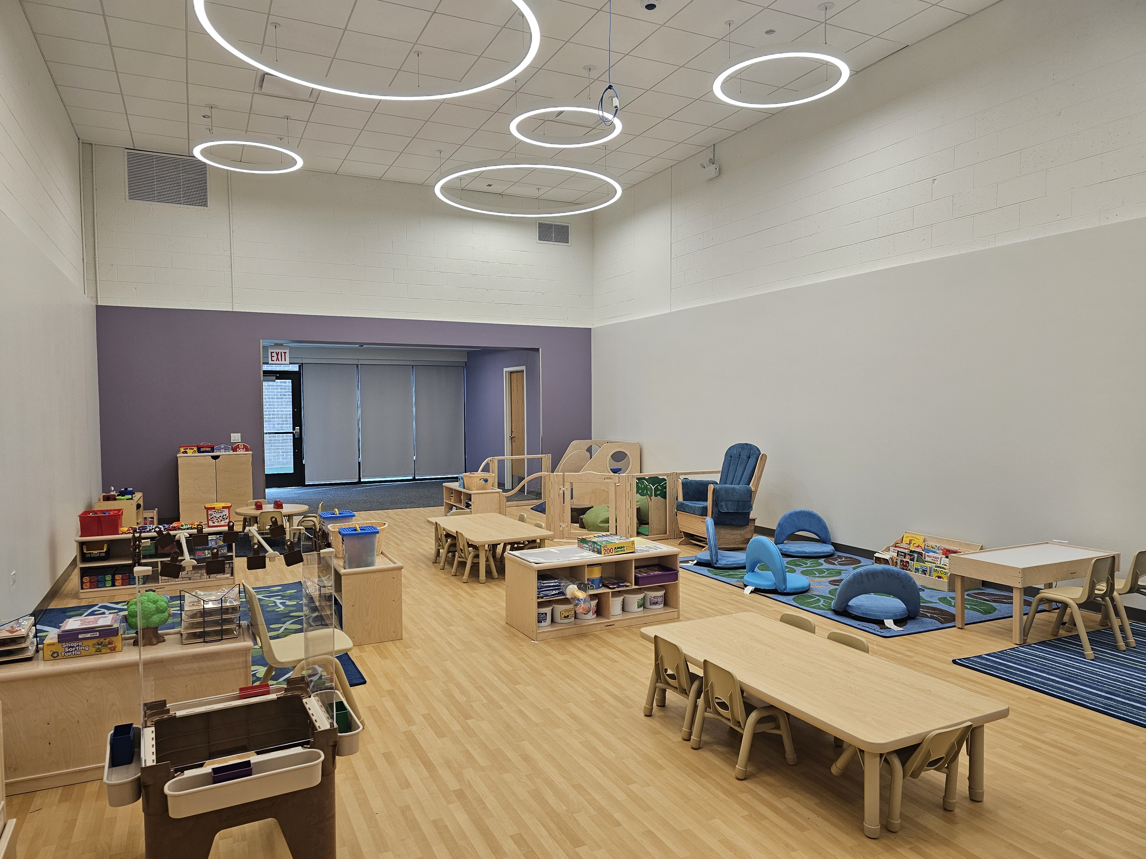 A toddler classroom with small, wooden tables and chairs, and a reading corner with books, a blue and brown area rug and floor cushions. There are multiple activity areas with easels for painting, and shelves full of toys and puzzles.