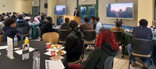 A man speaking at a podium to a large audience seated at round tables in a brightly lit room.