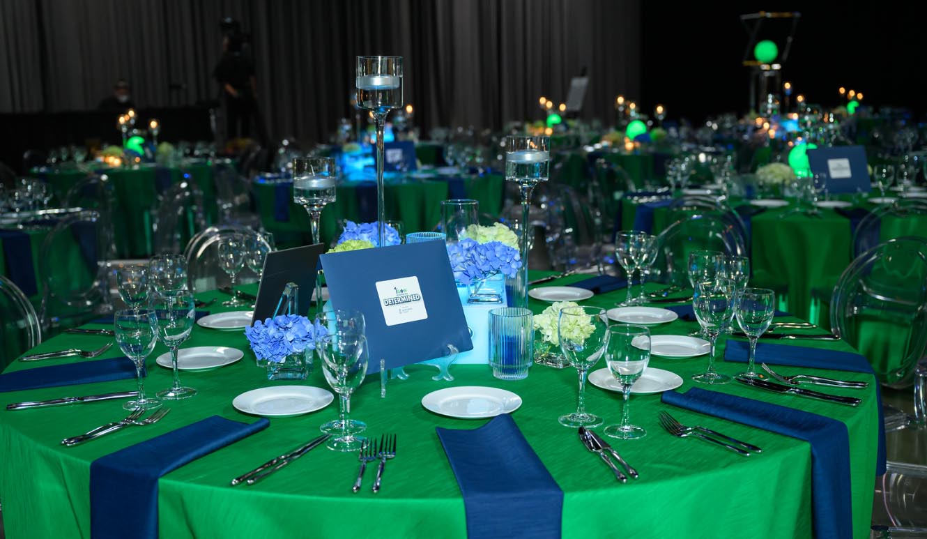 Banquet hall set for the 2025 1stGen Scholarship Gala, with round tables dressed in green linens, navy napkins, and floral centerpieces with candles. A place setting and event program reading “Determined” are visible in the foreground.