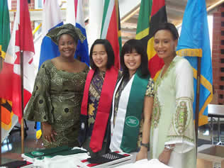 Four smiling women stand together at a university event, positioned in front of several international flags. The woman on the far left wears a green patterned traditional West African dress and headtie (gele). The two women in the center wear graduation stoles—one red and one green. The woman on the right wears a light green embroidered traditional dress. They are gathered behind a table with folded graduation regalia.