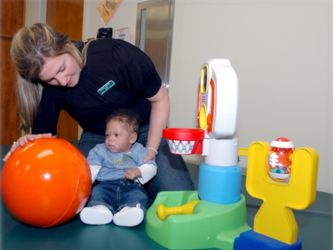 An occupational therapy student sits on the floor with an infant in her lap, while playing with a large, orange ball