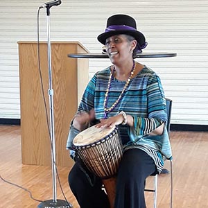 A woman sitting in a chair plays a drum and sings into a microphone.